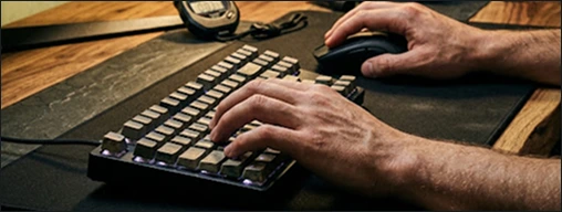 Mãos de um jogador utilizando um teclado mecânico iluminado e um mouse gamer sobre um mousepad em uma mesa de madeira.